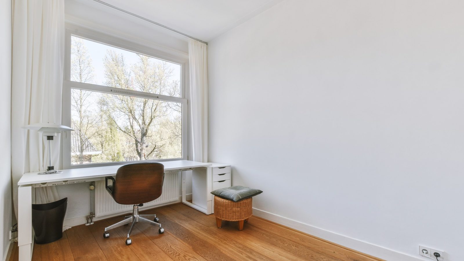 Helles Zimmer mit langem weißem Schreibtisch unter einem großen Fenster, davor ein Bürostuhl mit Holzschale, Blick auf Bäume
