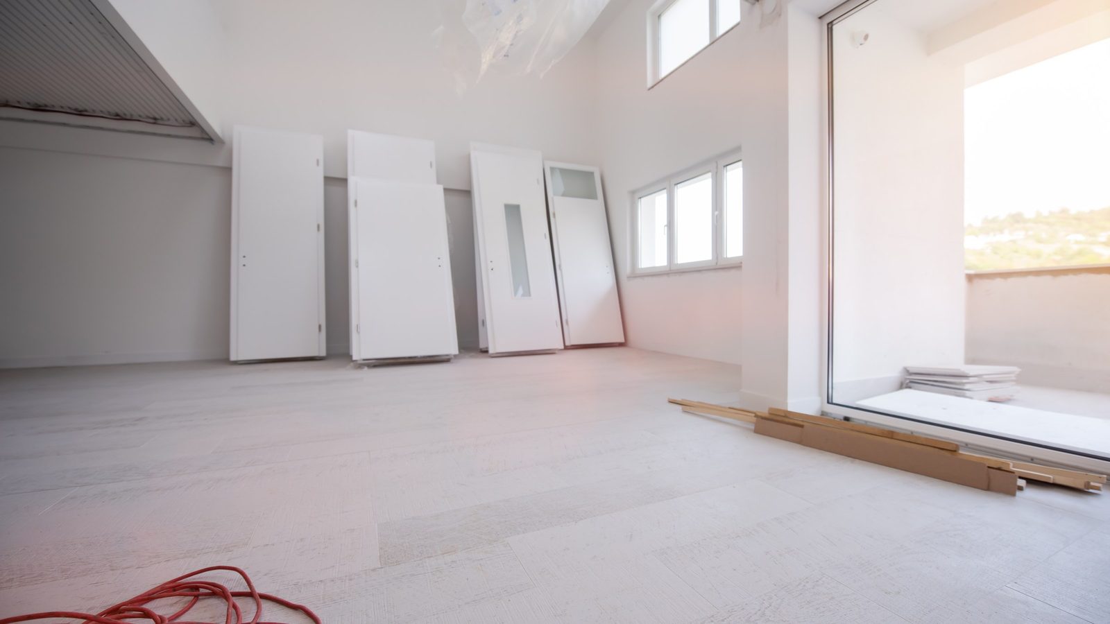 Heller Neubauraum mit grauem Boden, an die Wand gelehnten weißen Innentüren, einem Kabel auf dem Boden und einer offenen Balkontür