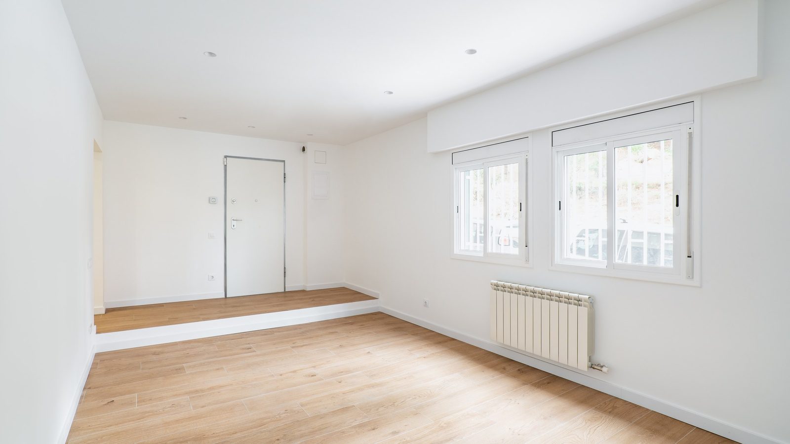 Leerer, frisch renovierter Raum mit hellem Holzboden, weißen Wänden, zwei Fenstern und einem weißen Heizkörper