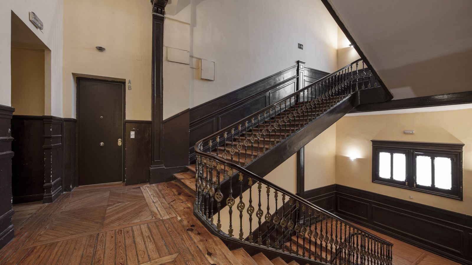 Treppenhaus in einem Altbau mit Dielenboden im Fischgrätmuster, dunkler Holzvertäfelung, verziertem Eisengeländer und einer geschlossenen Wohnungstür