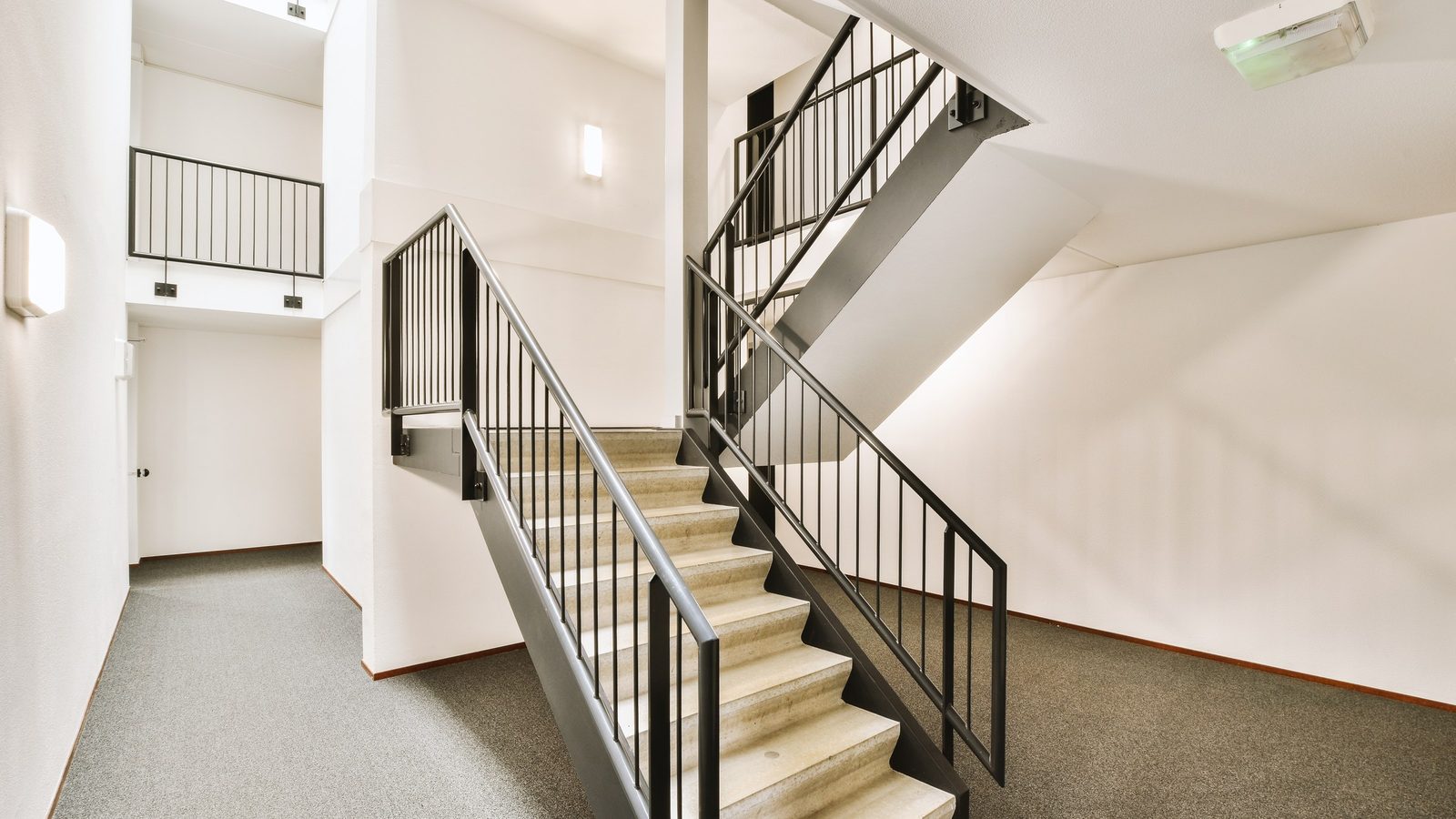 Helles Treppenhaus mit weißen Wänden, grauem Teppichboden und einer Steintreppe mit schwarzem Metallgeländer