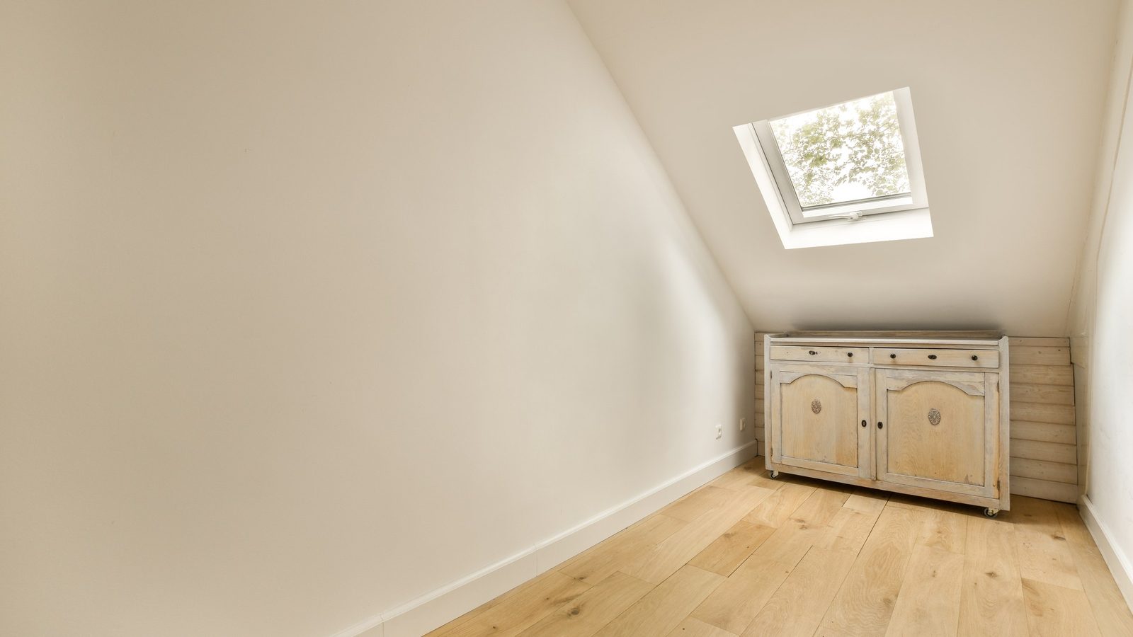 Schmales Zimmer unter der Dachschräge mit Holzdielen, einem offenen Dachfenster und einer hellen Kommode an der Stirnseite