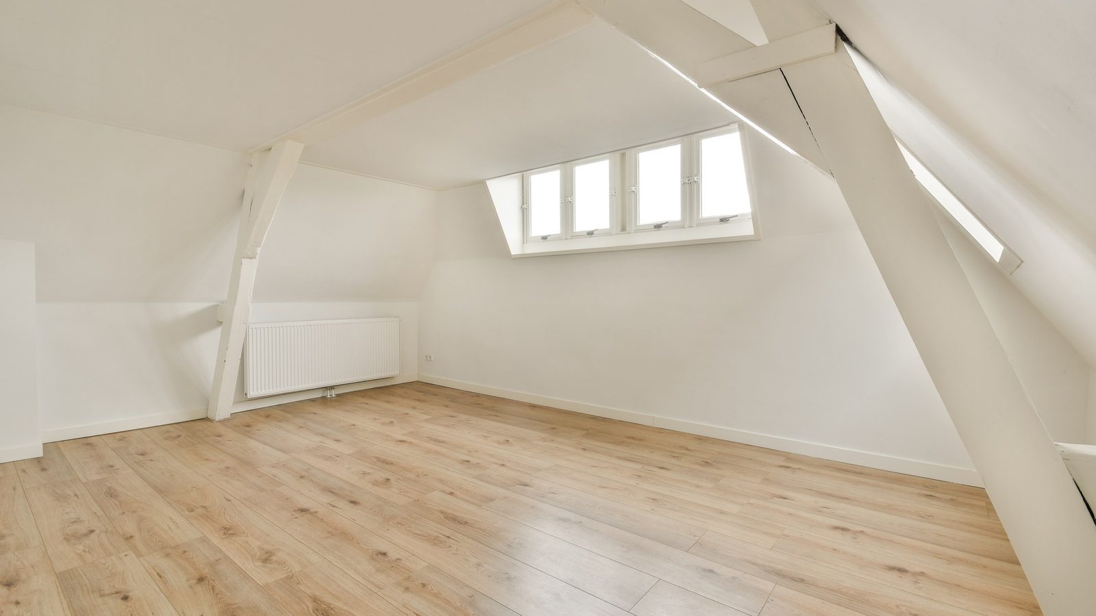 Leeres Dachzimmer mit hellem Laminatboden, weiß gestrichenen Dachbalken, einer Gaube mit vier Fenstern und einem Heizkörper an der Wand