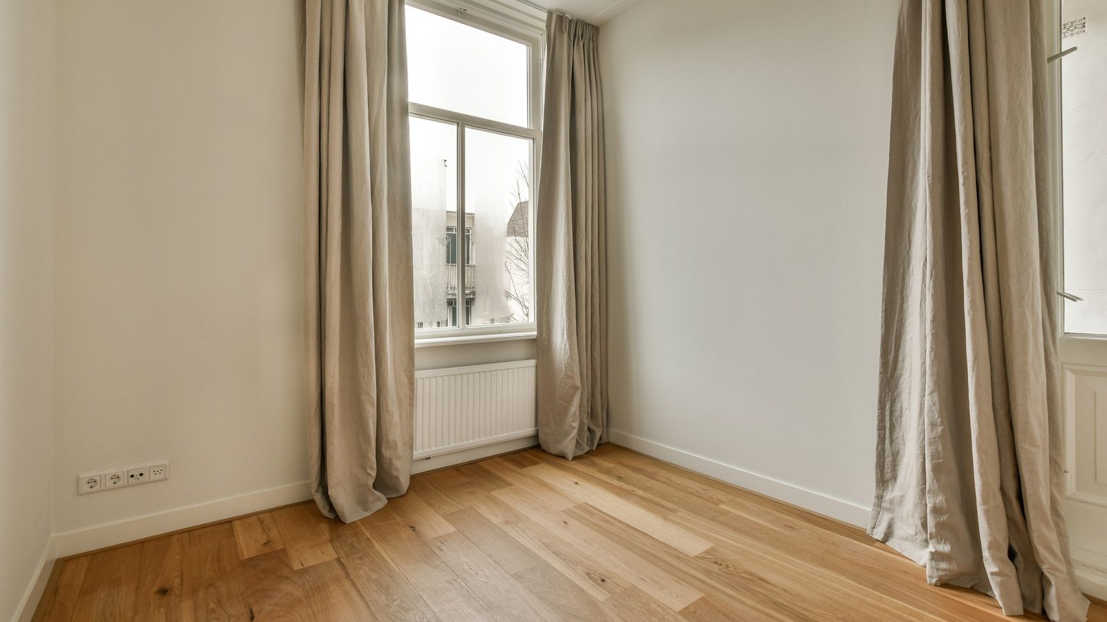 Leeres Zimmer mit Holzdielen, hohen beigen Vorhängen an einem Sprossenfenster und einem weißen Heizkörper