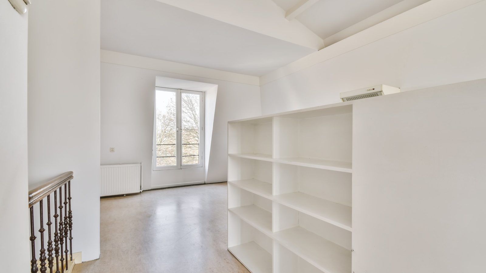 Leerer Raum im Dachgeschoss mit einem weißen offenen Regal, einem Treppengeländer links und einem Sprossenfenster mit Blick auf Bäume