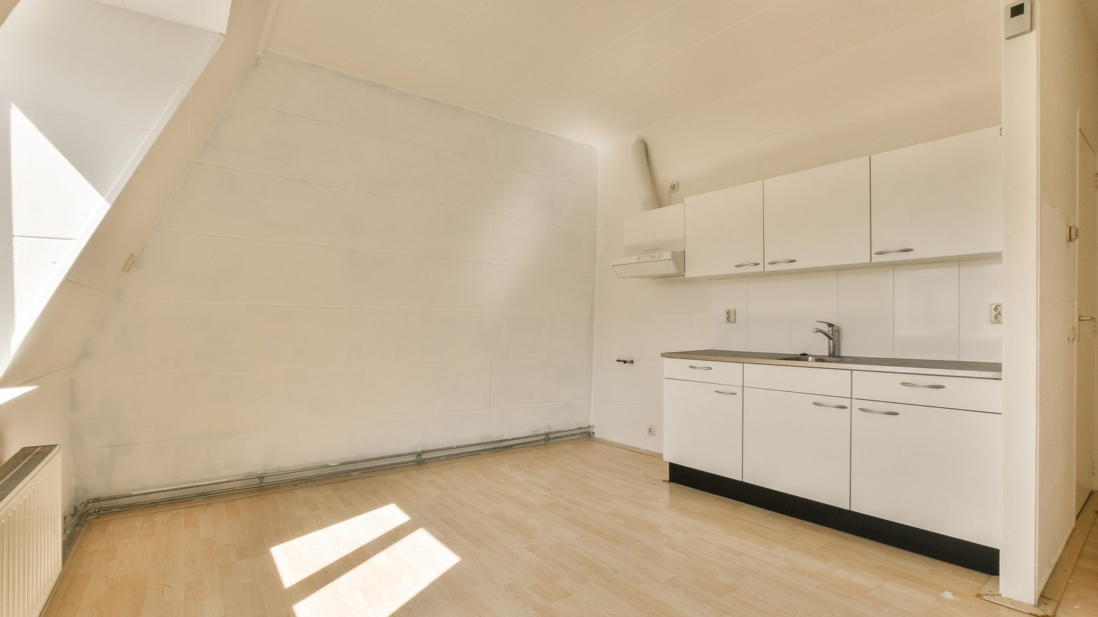 Leere Dachgeschosswohnung mit weißer Küchenzeile, hellem Holzboden und Sonnenlicht durch ein Dachfenster