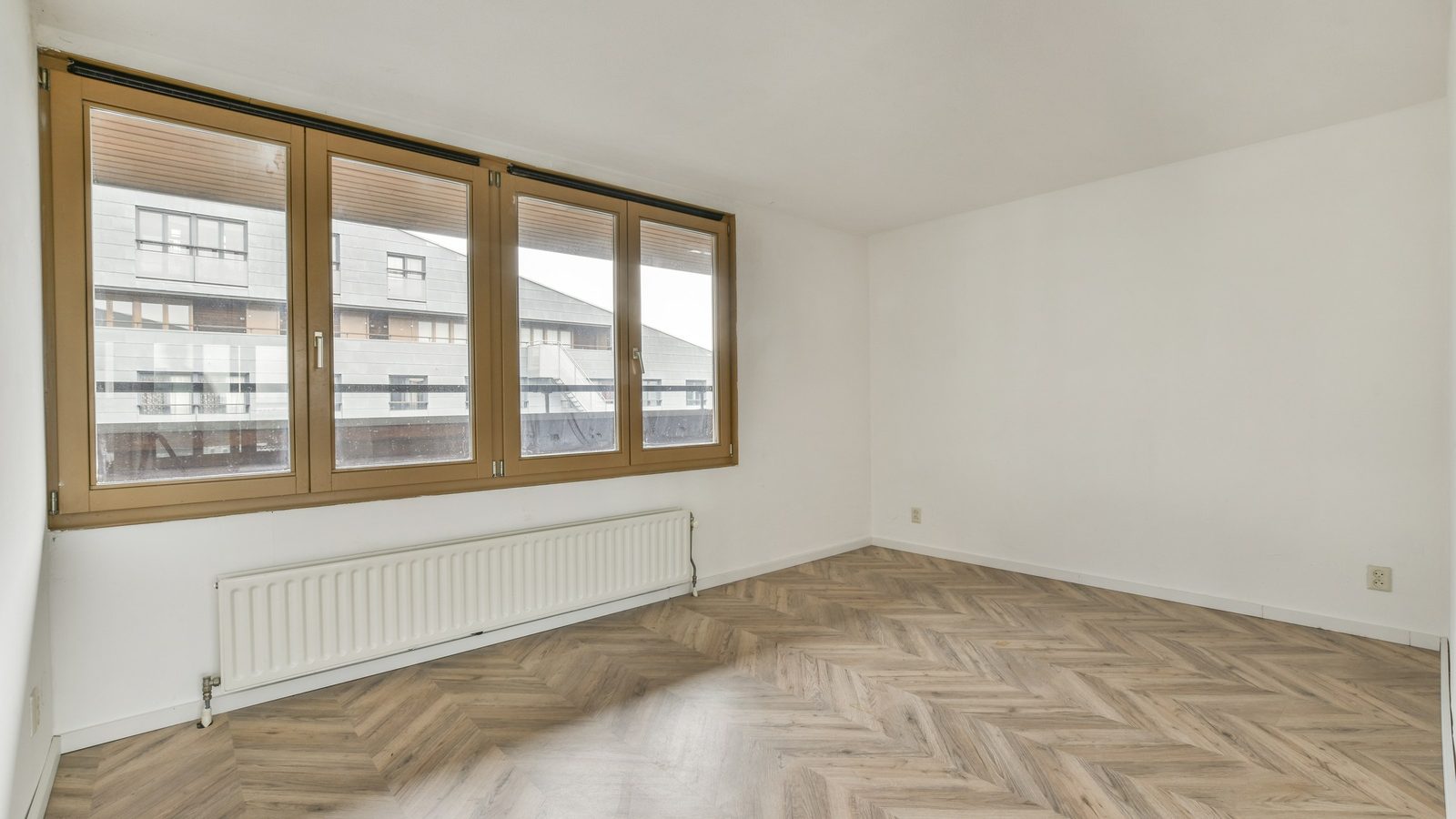 Leeres Zimmer mit Fischgrätparkett, weißem Heizkörper unter einem breiten Holzfenster mit Blick auf ein Wohnhaus
