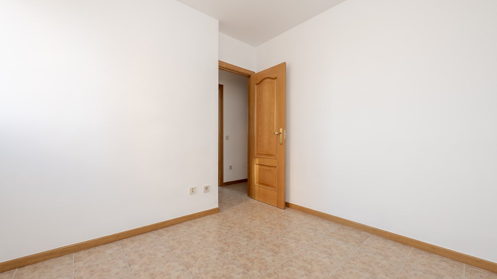 Leeres Zimmer mit weißen Wänden, Fliesenboden und einer halb geöffneten Holztür zum Flur