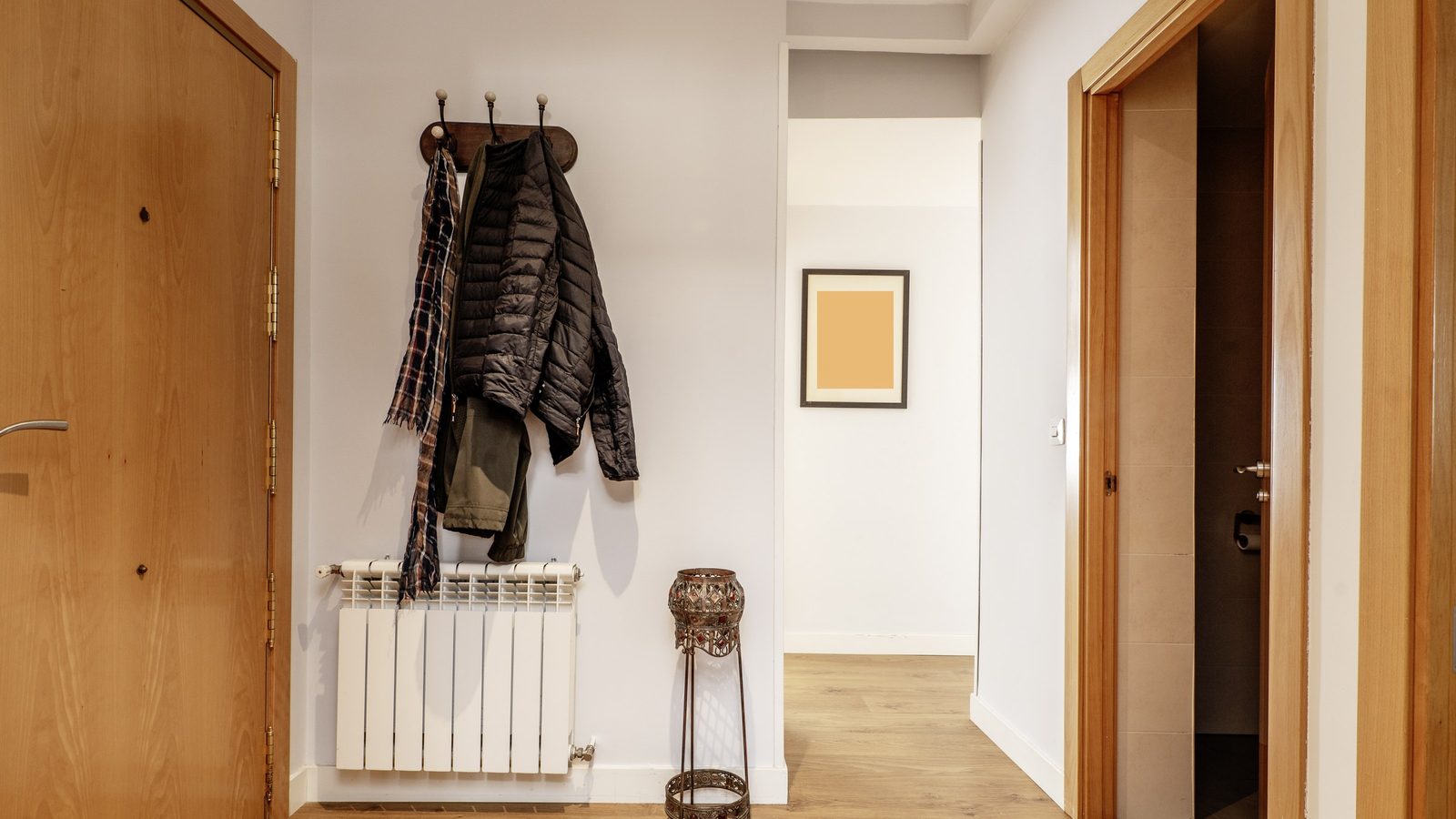 Wohnungsflur mit Holztür, einer Hakenleiste mit Jacke und Schal über einem weißen Heizkörper und Blick in einen hellen Gang